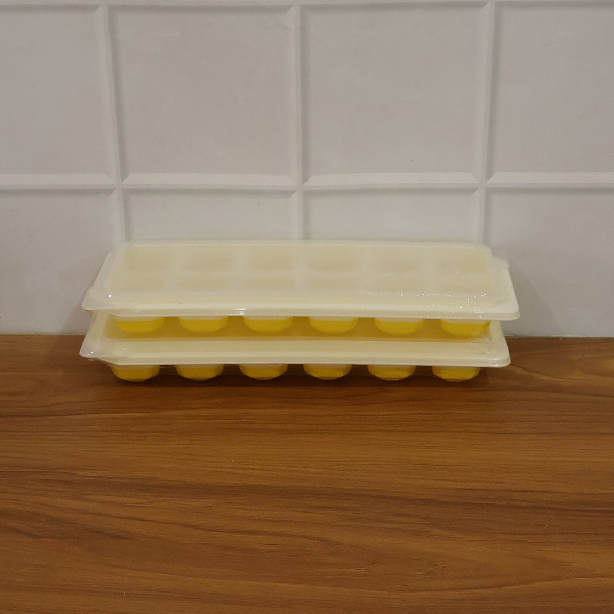 Two yellow silicone ice cube trays with clear lids are stacked on a wooden surface against a white tiled wall.