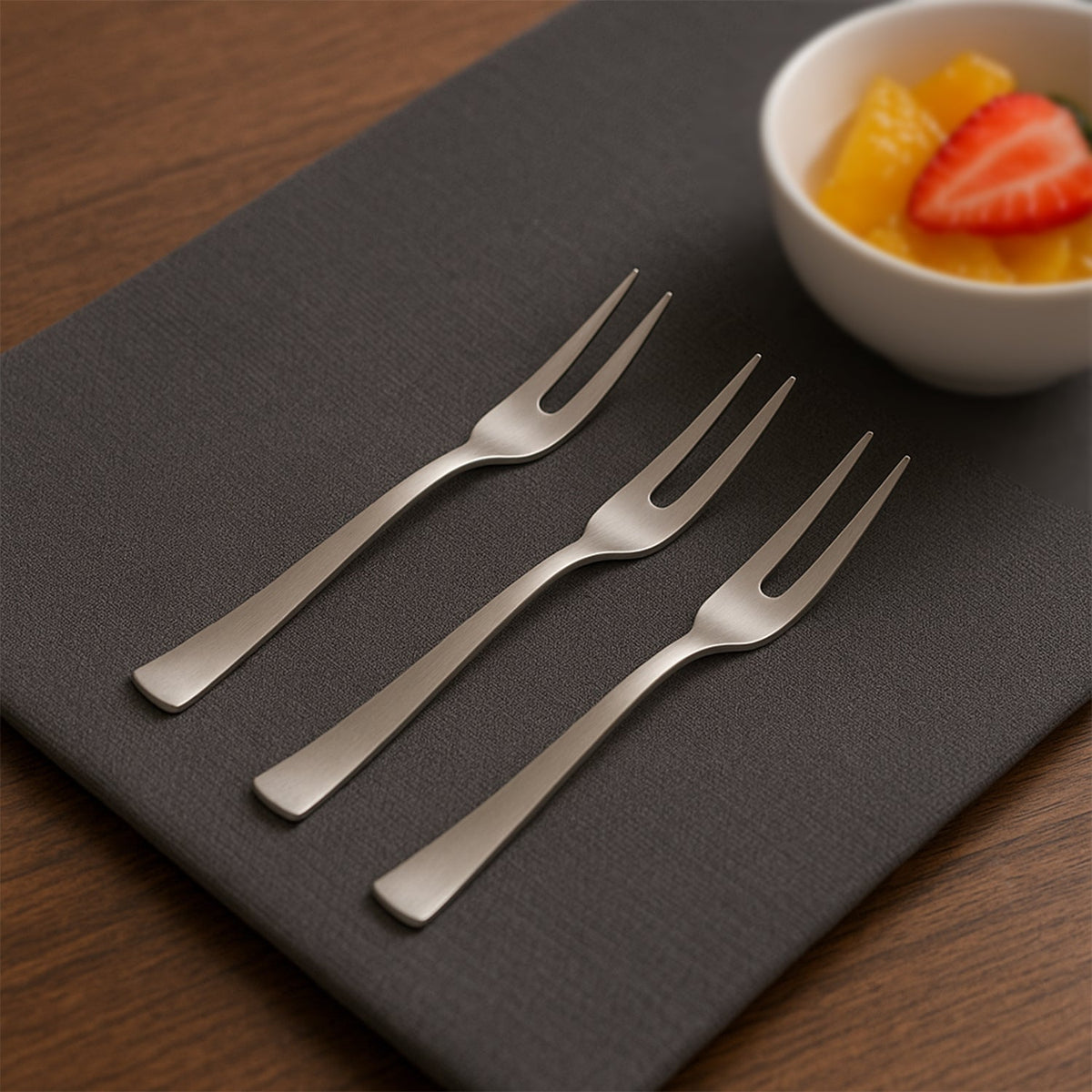 Three silver stainless steel fruit forks on a dark gray placemat next to a white bowl of fruit salad.