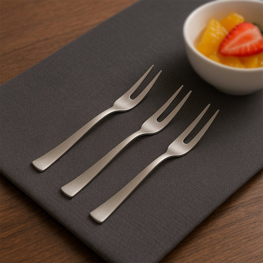 Three silver stainless steel fruit forks on a dark gray placemat next to a white bowl of fruit salad.
