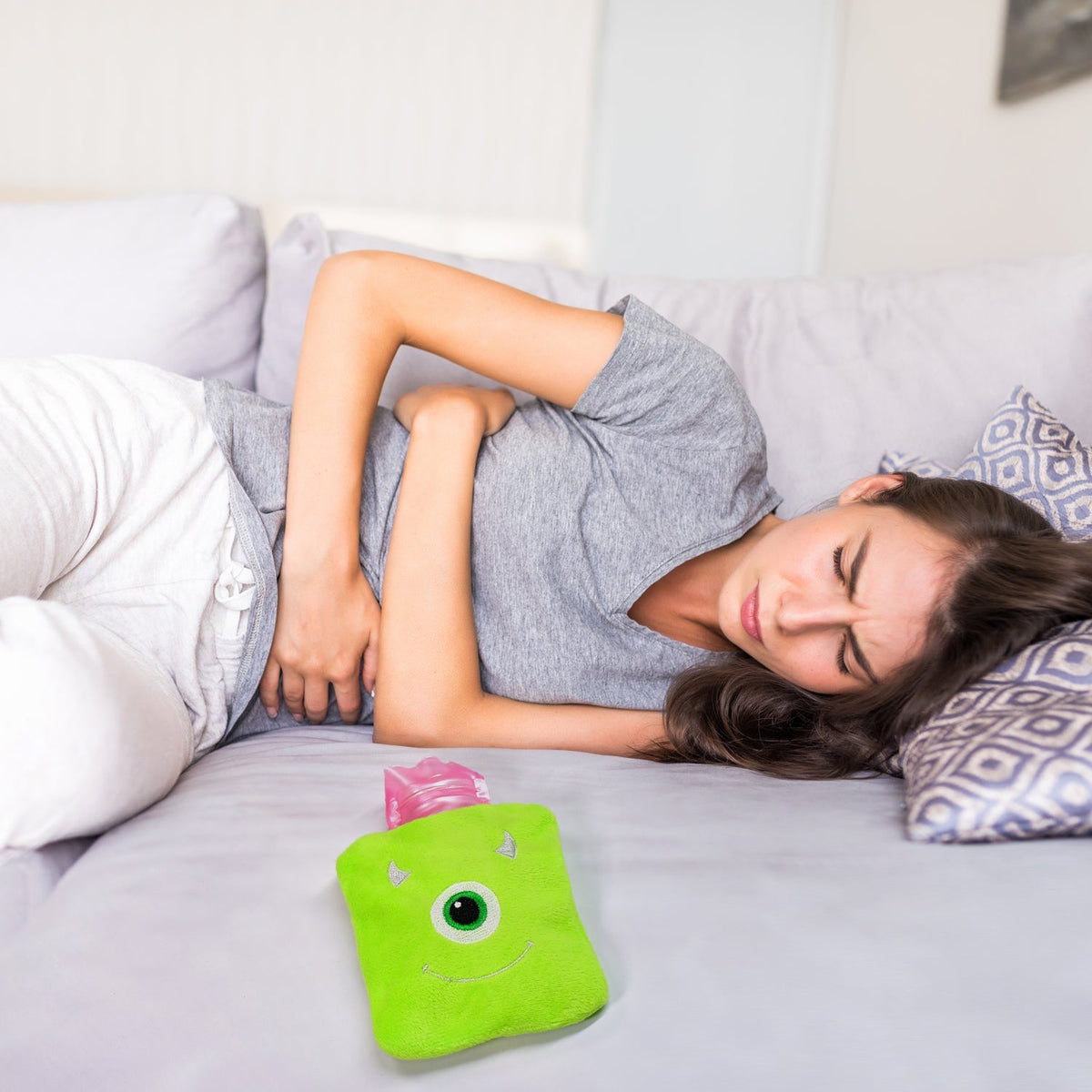 A woman lies on a gray couch, clutching her stomach in discomfort, with the green monster hot water bag nearby.