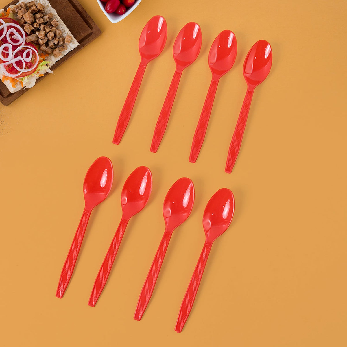 Eight red plastic spoons with textured handles arranged on a vibrant orange background with food nearby.