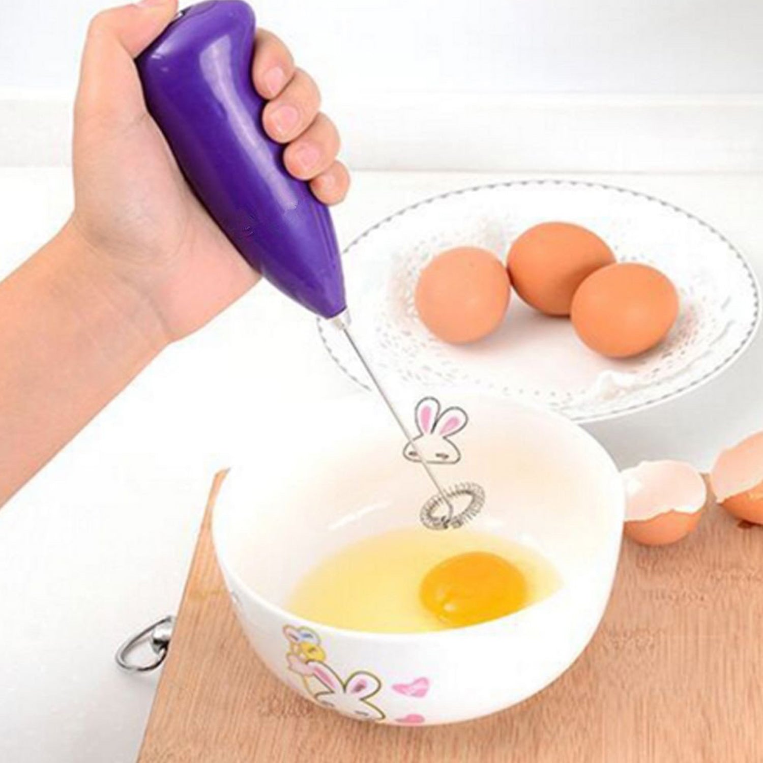 A hand holds a purple frother with a rabbit-shaped whisk, mixing a cracked egg in a white bowl.