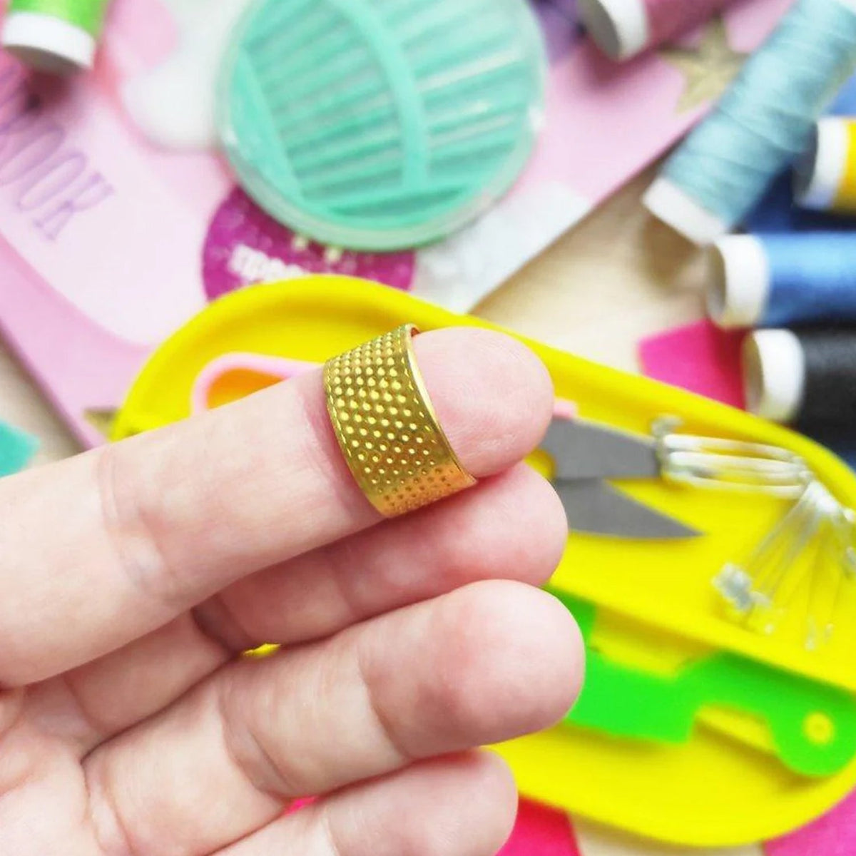 A close-up of a hand wearing a gold-colored thimble, with other sewing tools blurred in the background.