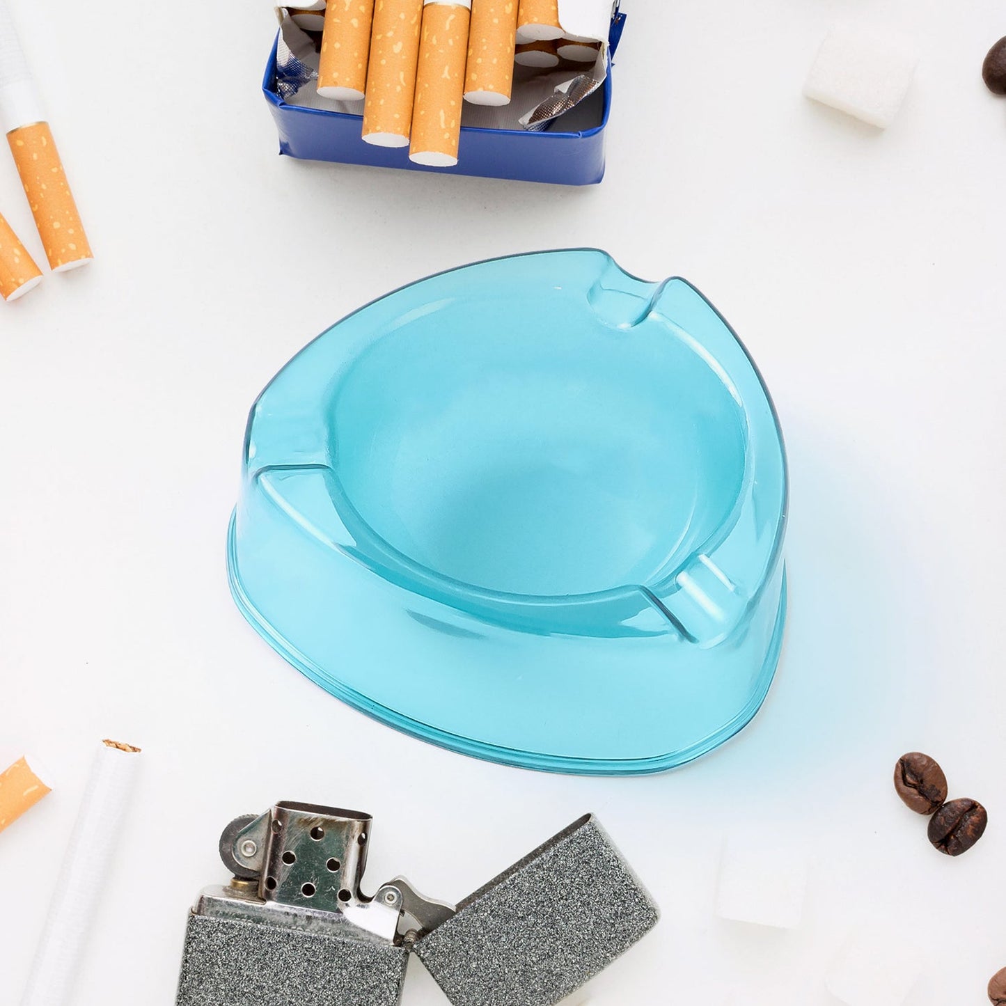 A clear transparent plastic ashtray is surrounded by several loose cigarettes on a plain white surface.
