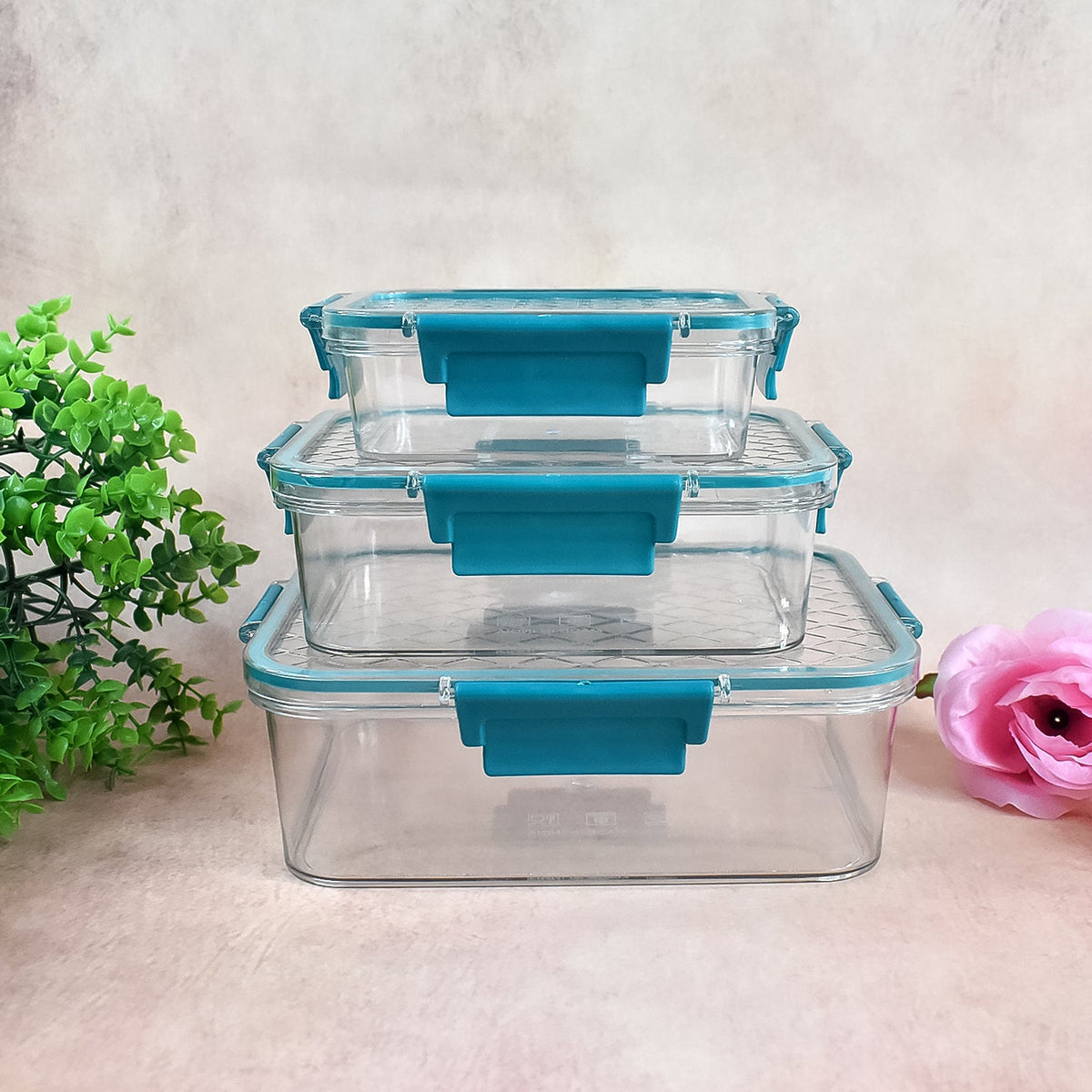 Three clear rectangular food containers with teal locking lids are stacked, with green foliage and a pink flower.
