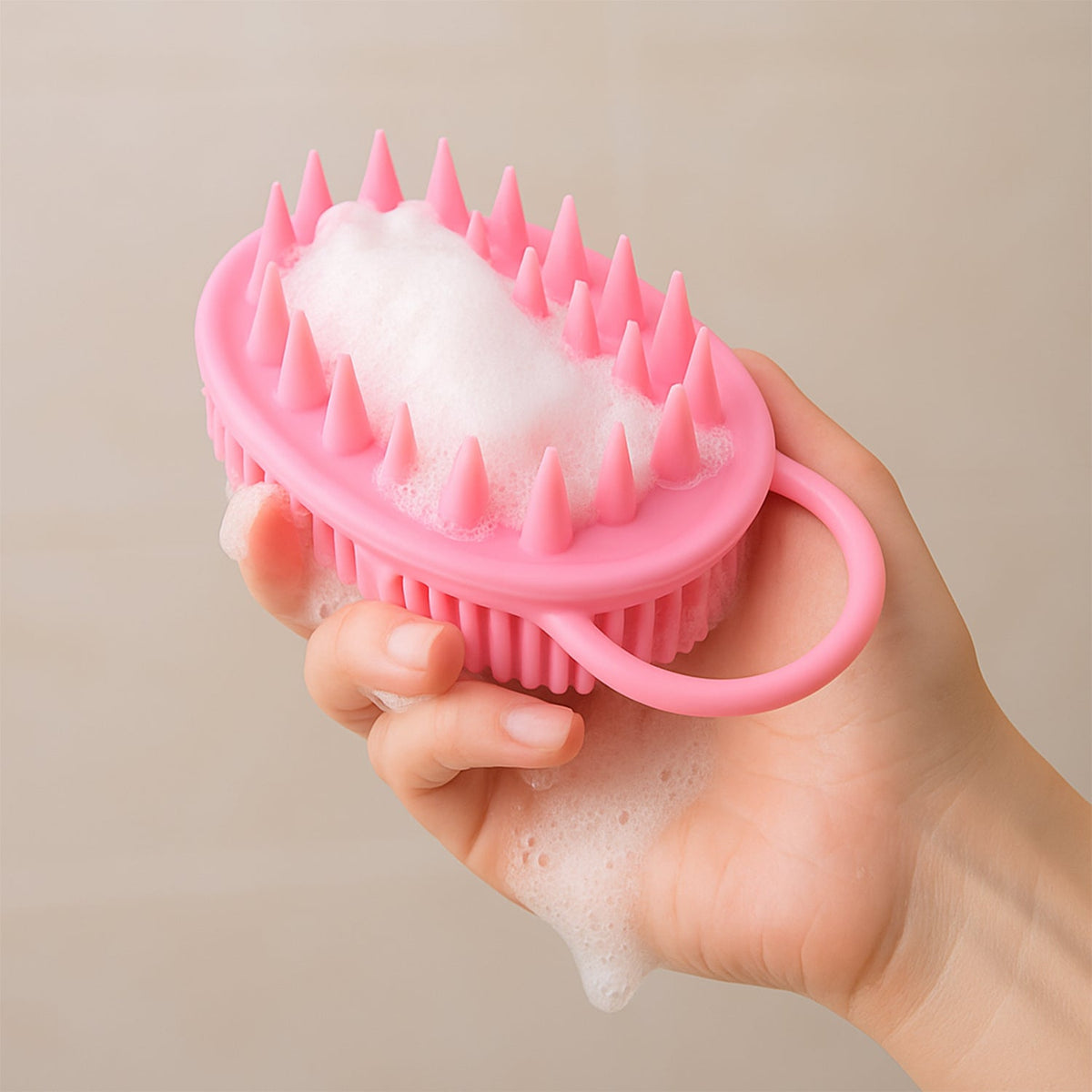 A hand holds a pink silicone body brush covered in white soap suds, with water dripping from the bristles.