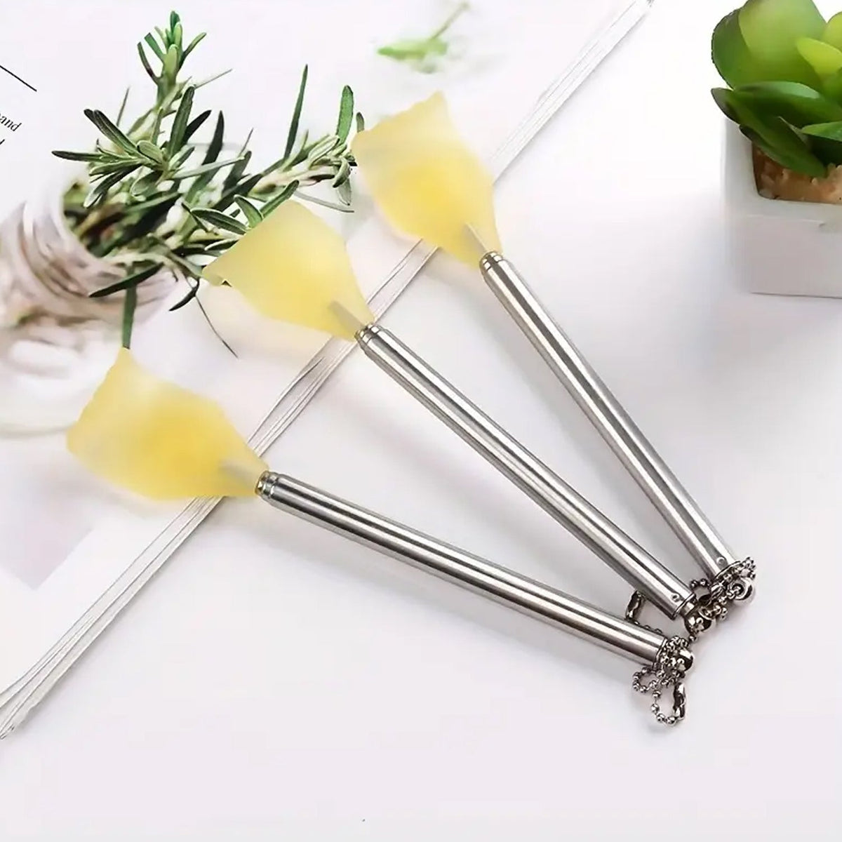 Three silver telescoping back scratchers with yellow hand-shaped heads lying on a white surface with a plant in the