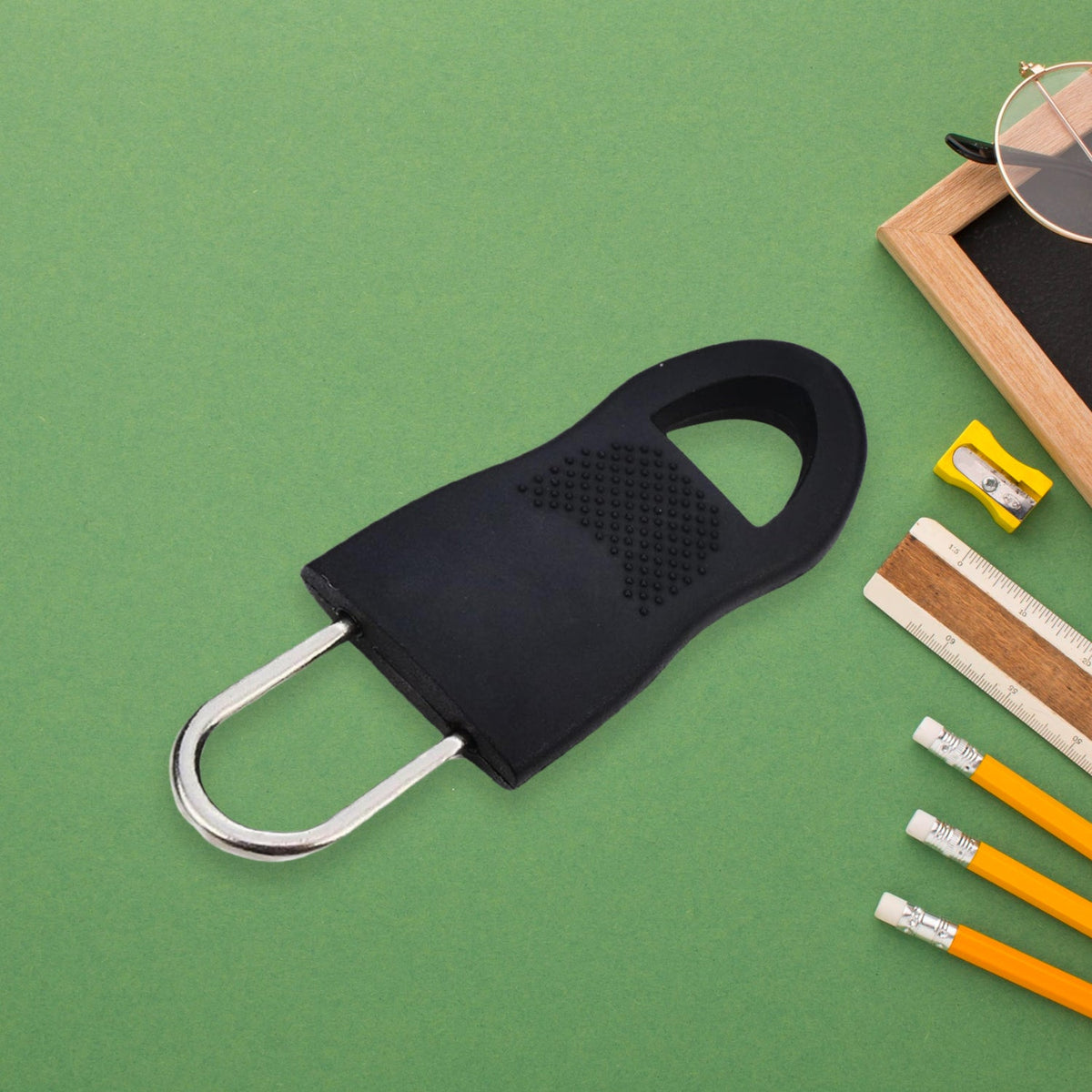A black rubber zipper pull tab with a silver metal loop is on a green surface with pencils and a ruler.