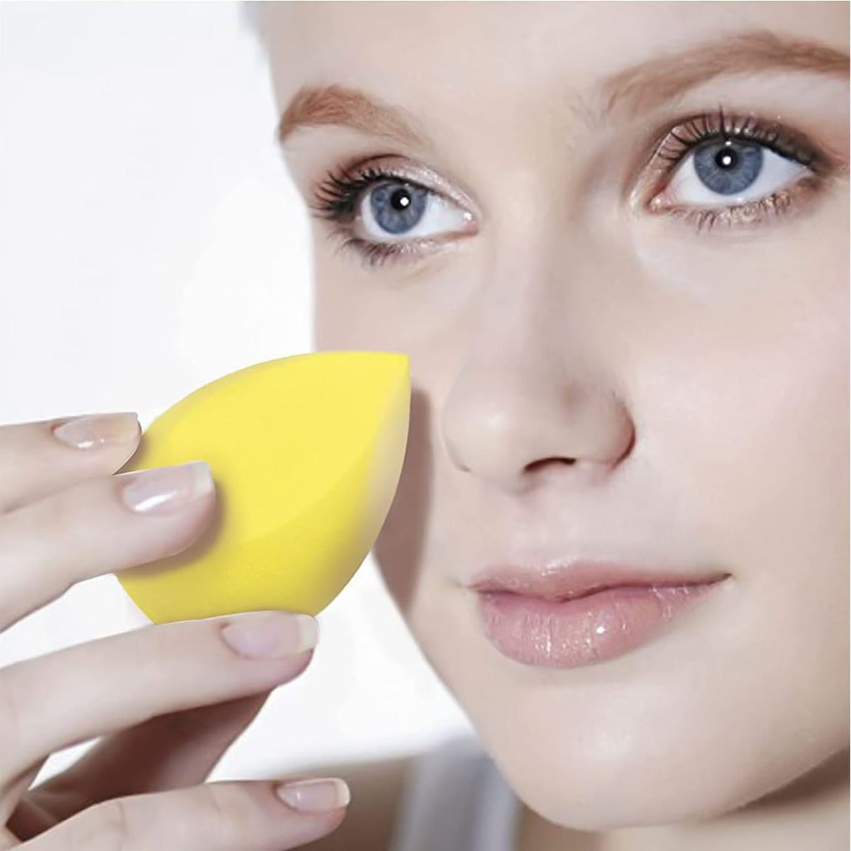 A woman with blue eyes holds a bright yellow teardrop makeup sponge near her cheek.