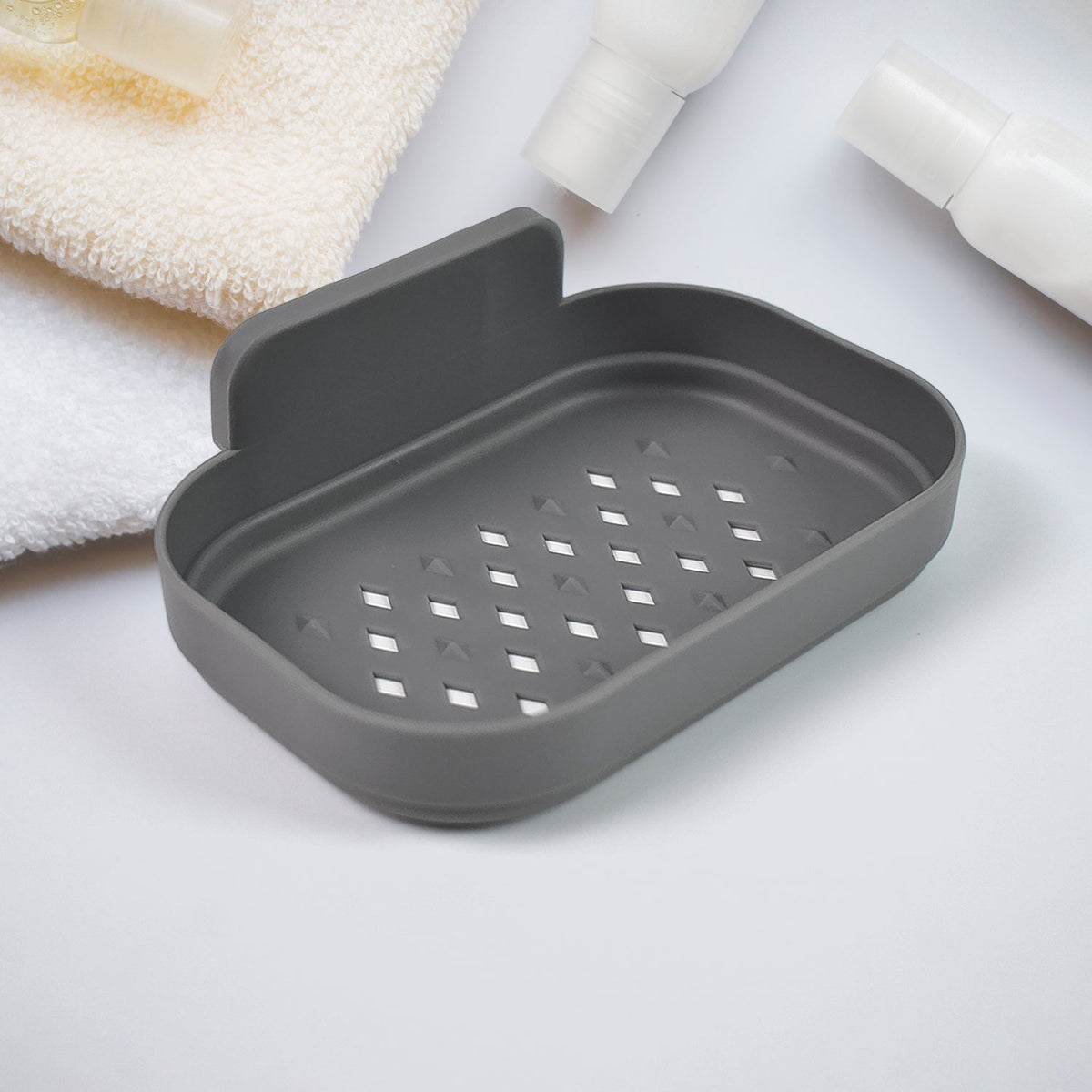 A gray self-adhesive soap dish with drainage holes is shown on a white surface with towels and bottles.