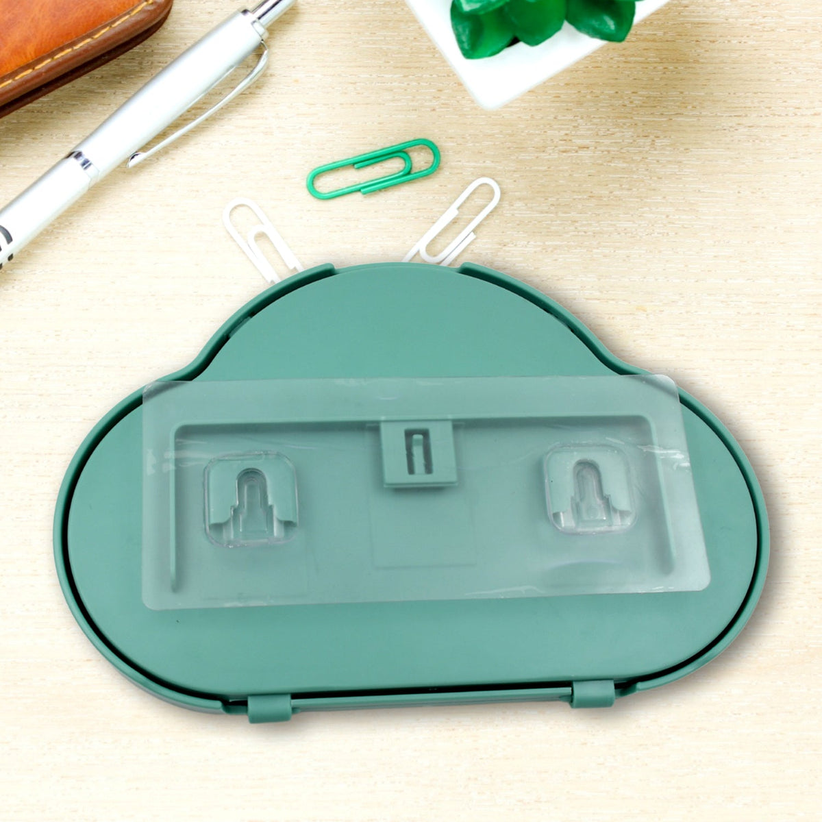 The back of a teal cloud-shaped phone holder with clear adhesive strips and hooks, on a desk.