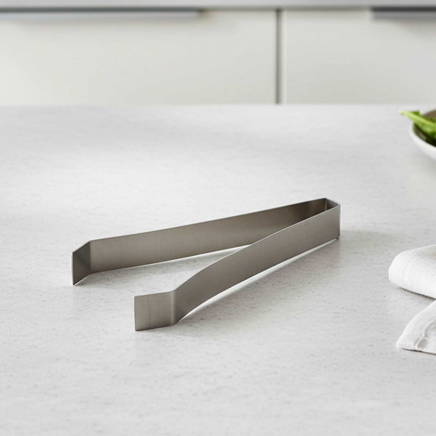 A single silver stainless steel tweezer lies on a white kitchen counter with a blurred background.