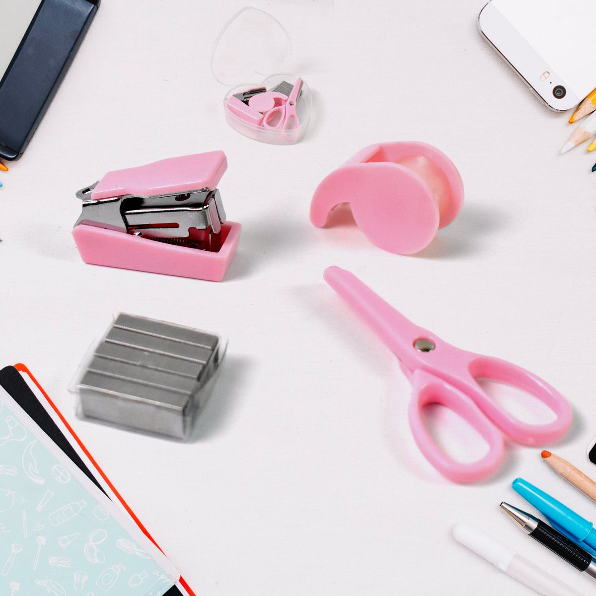 A pink mini stapler, tape dispenser, scissors, and a box of staples arranged on a white surface with other office supplies.