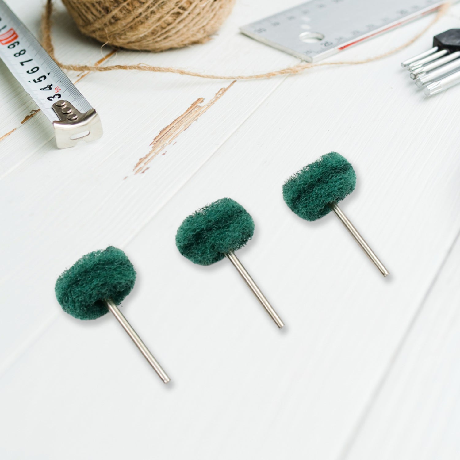 Three green polishing wheels with metal shanks arranged diagonally on a white wooden surface, with various tools in the