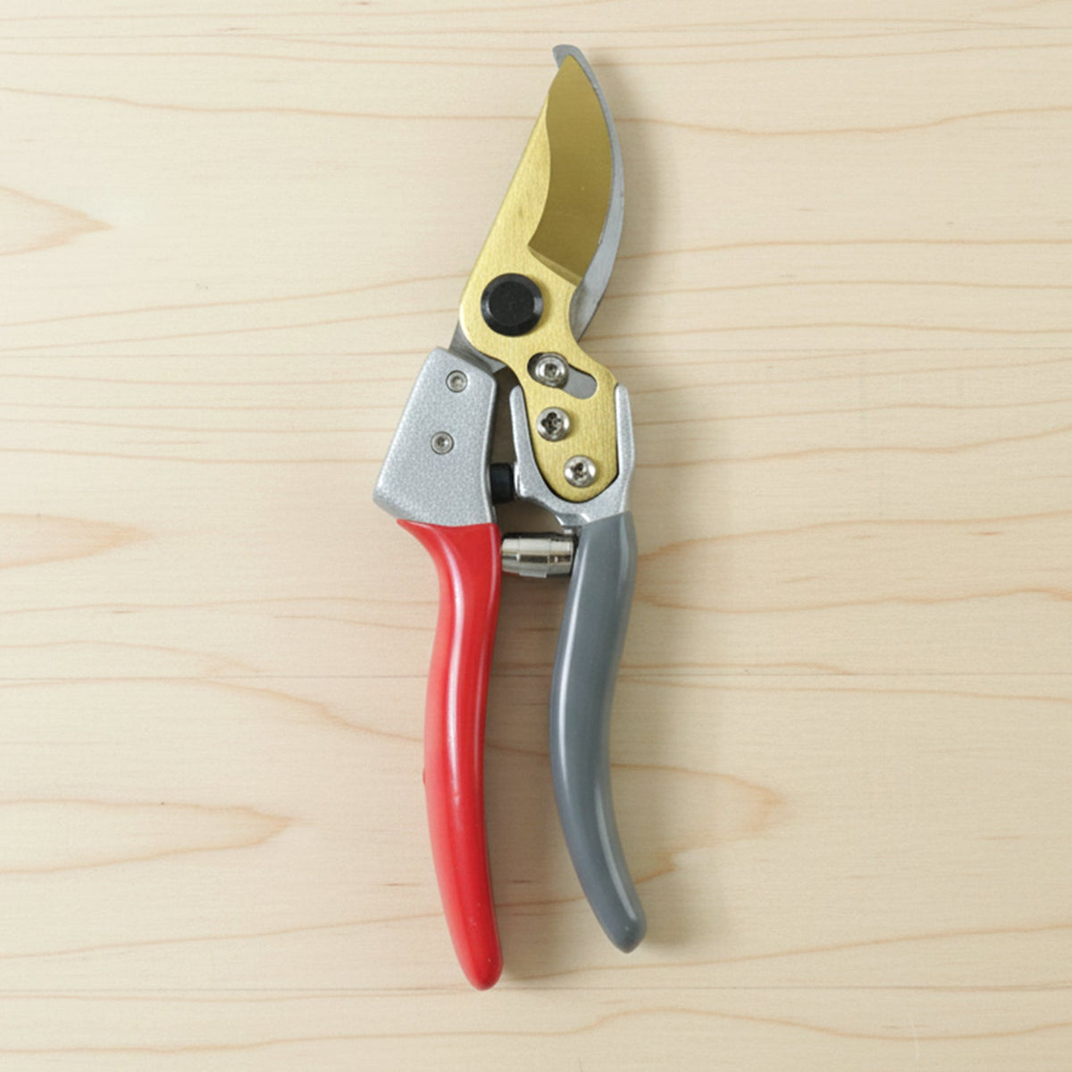 Red and grey garden shears with gold blades lying flat on a light wooden surface.