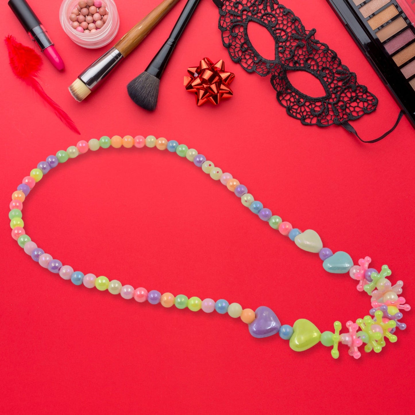 A colorful beaded necklace with heart and star-shaped beads is laid on a red surface surrounded by various makeup products.