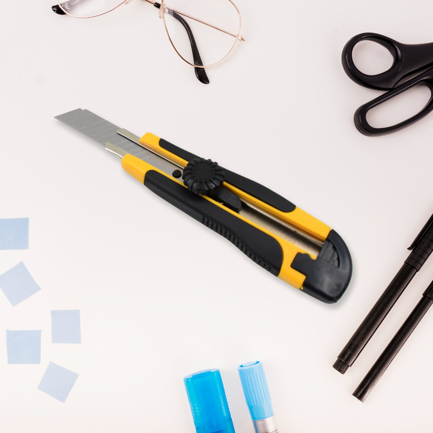 A black and yellow utility knife is centrally placed on a white surface with glasses, scissors, and pens around it.