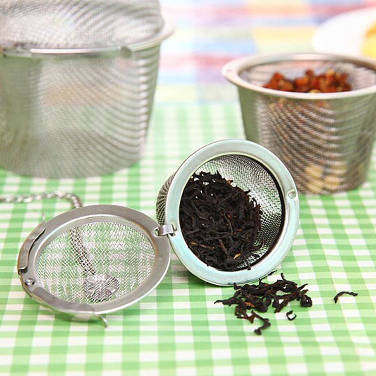 An open stainless steel tea infuser filled with black tea leaves, with more loose tea and other infusers on a green