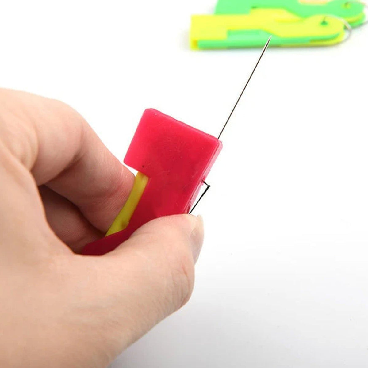 A hand holding a red and yellow needle threader with a needle protruding from the top.