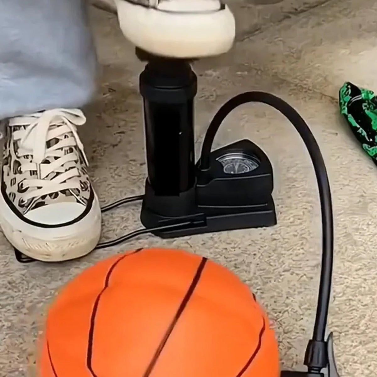 A person's feet in sneakers operating a black foot pump to inflate an orange basketball.