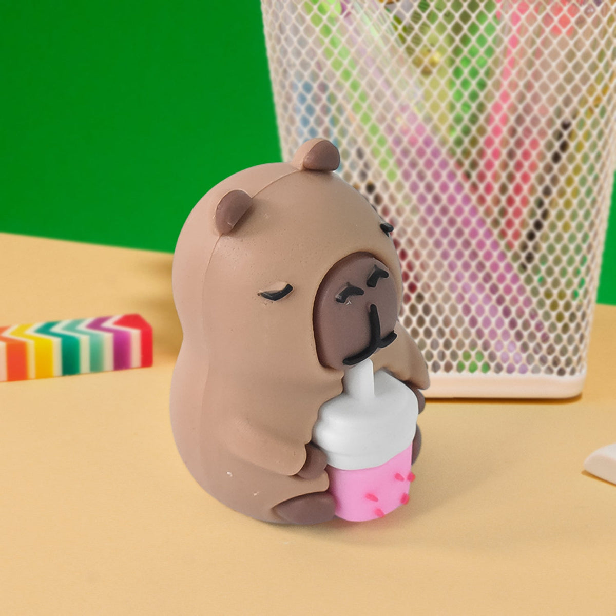 A capybara-shaped pencil sharpener with a pink drink sits on a yellow surface next to a white mesh pen holder.