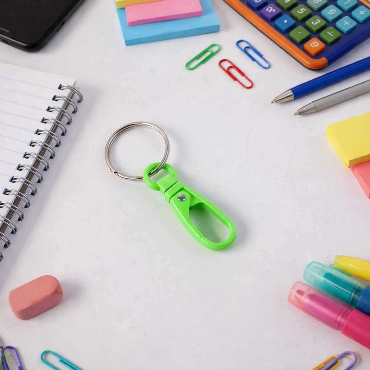 A green plastic carabiner keychain with a silver ring on a white desk surrounded by office supplies.