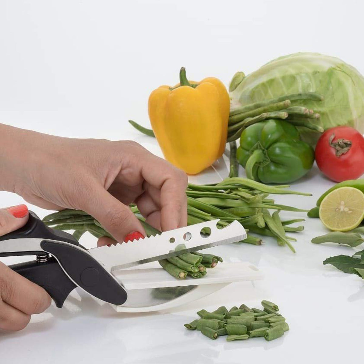 A person's hands use a clever cutter to chop green beans on a white surface, surrounded by various vegetables.