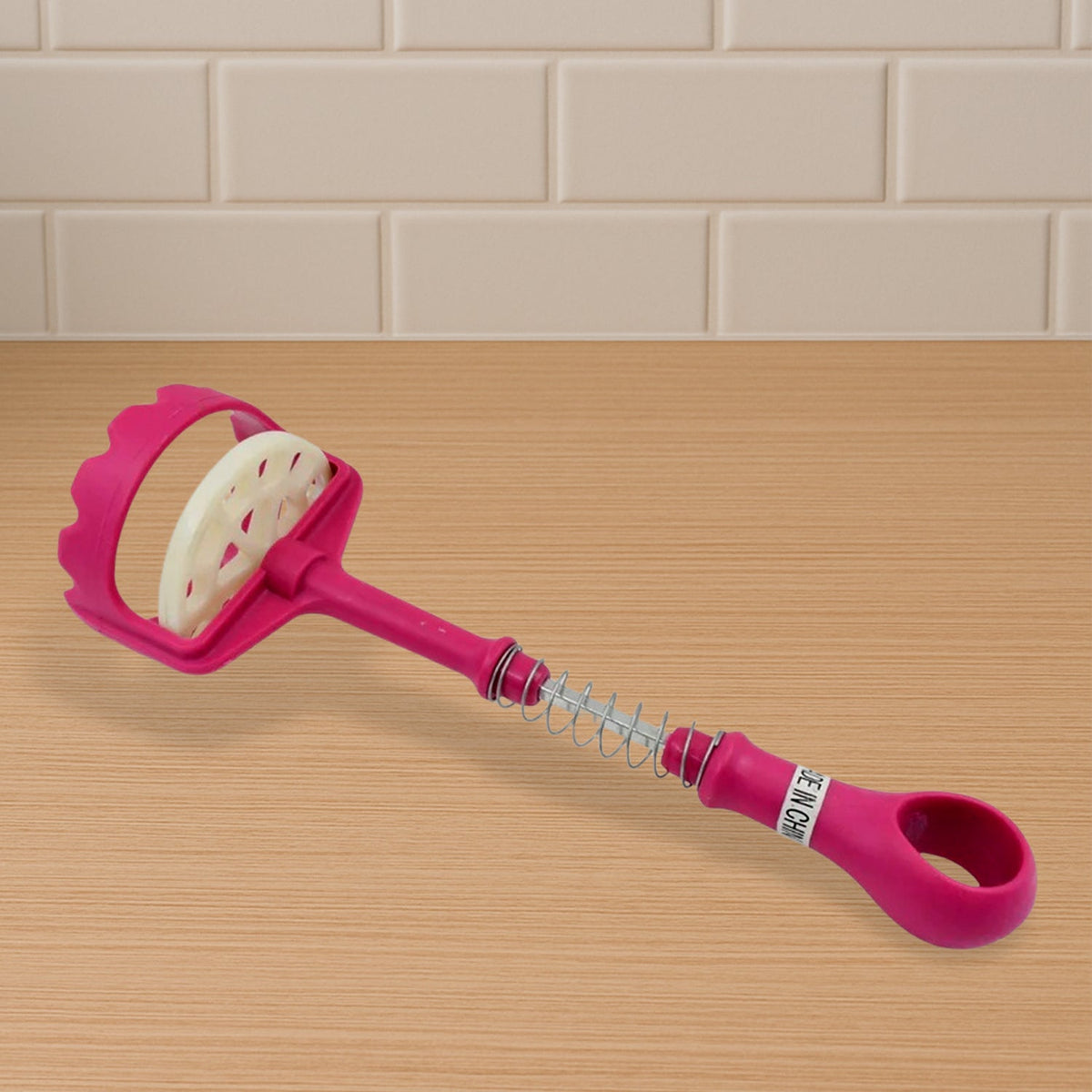 A pink and white plastic potato masher lies on a wooden surface, showing its spring mechanism.