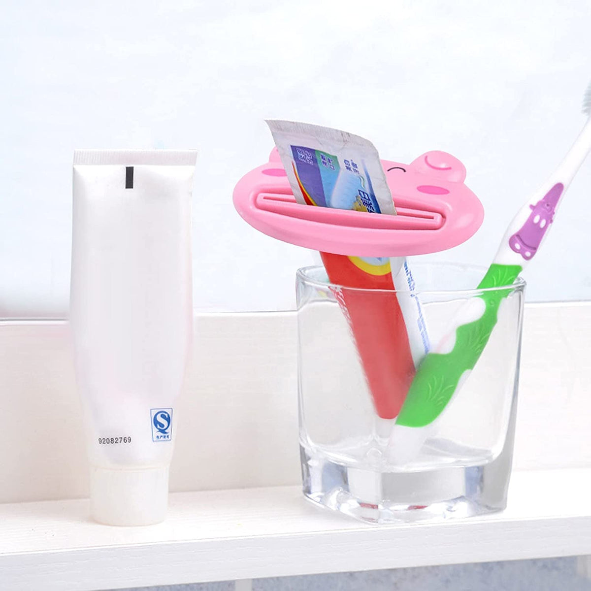 A pink pig-shaped toothpaste squeezer holding a red and white toothpaste tube upright in a clear glass next to a white tube
