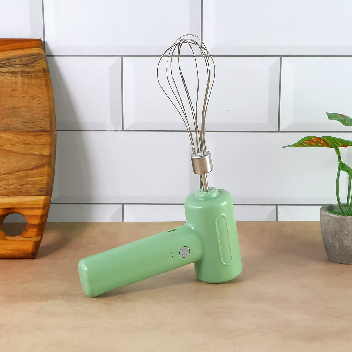 Light green cordless electric whisk standing upright on a brown counter, next to a wooden cutting board and a plant.