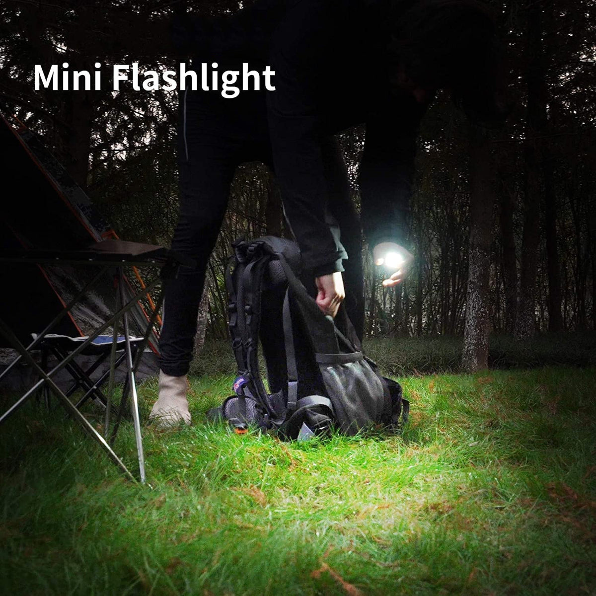 A person in a dark forest setting, holding a small, bright flashlight that illuminates the ground and their backpack.