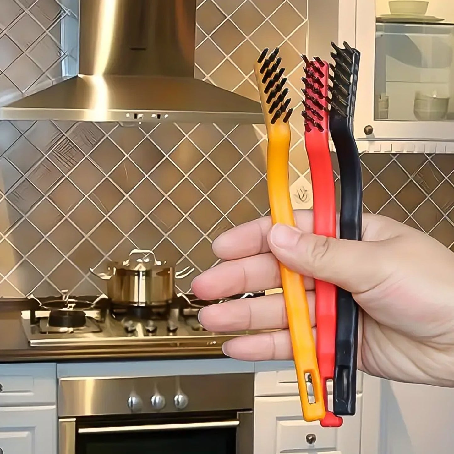 A hand holds three wire brushes (yellow, red, black) in a kitchen setting with a stove and range hood in the background.