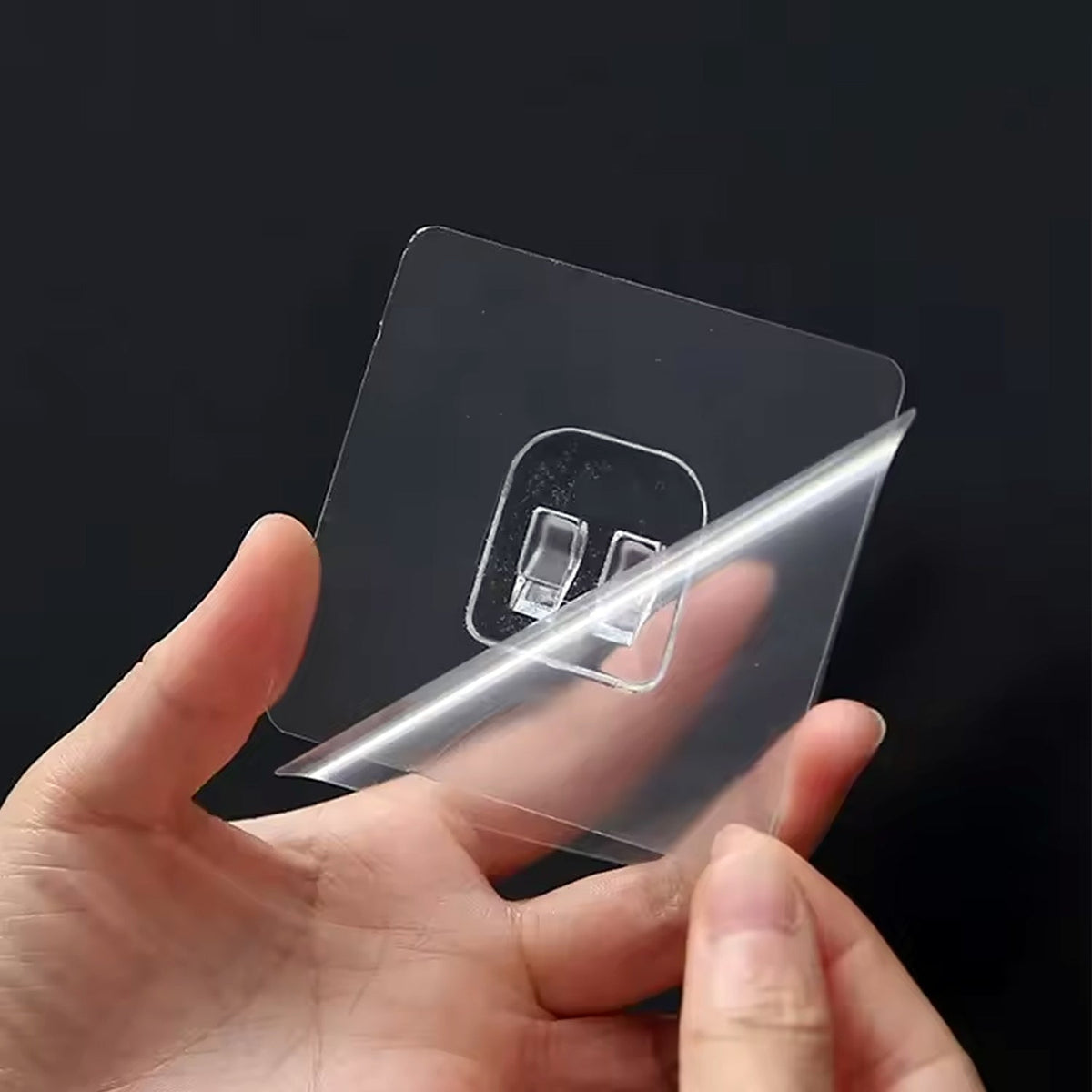 Hands peel the protective film off a clear self-adhesive wall hook with two prongs against a dark background.