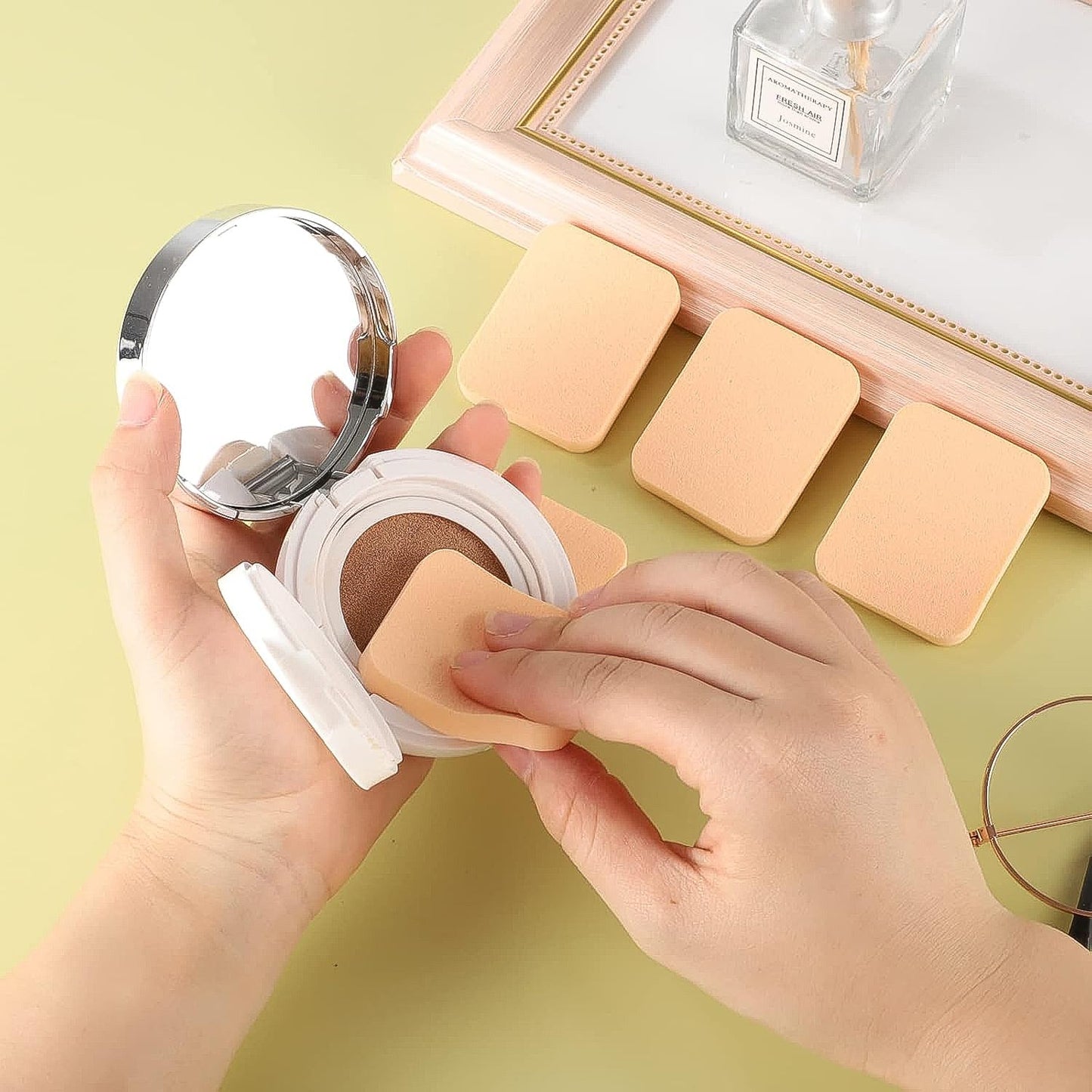 Hands hold a compact cushion foundation with a beige square sponge, with three additional sponges on a light green surface.
