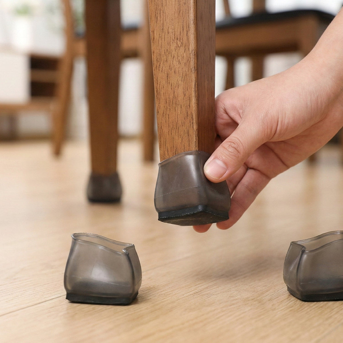 A hand places a dark grey silicone cap onto a wooden chair leg, with two other caps on the floor.
