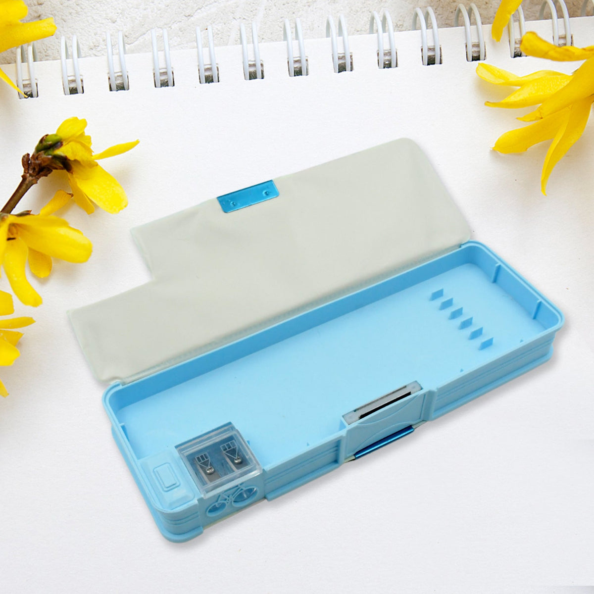 Open blue pencil case showing the empty main compartment and built-in sharpener, with yellow flowers nearby.