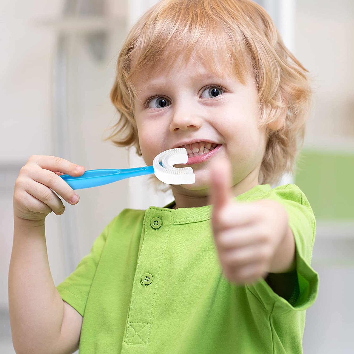 A smiling boy in a green shirt holds a blue U-shaped toothbrush in his mouth and gives a thumbs up.