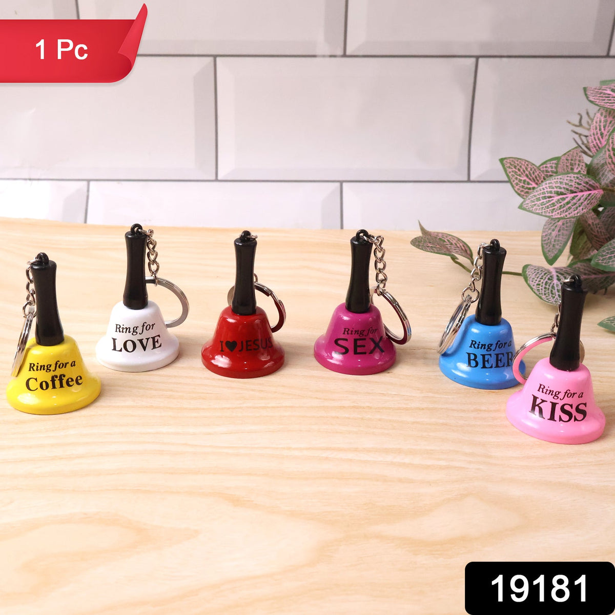Six mini hand bell keychains in different colors with phrases, arranged on a wooden surface against a white tiled background.
