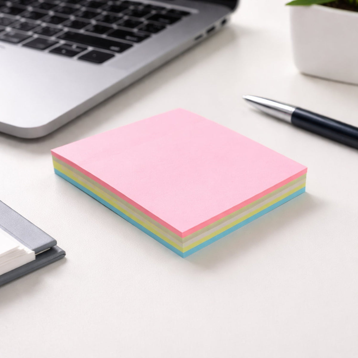 A stack of pink, yellow, and blue sticky notes on a white desk next to a laptop and a black pen.