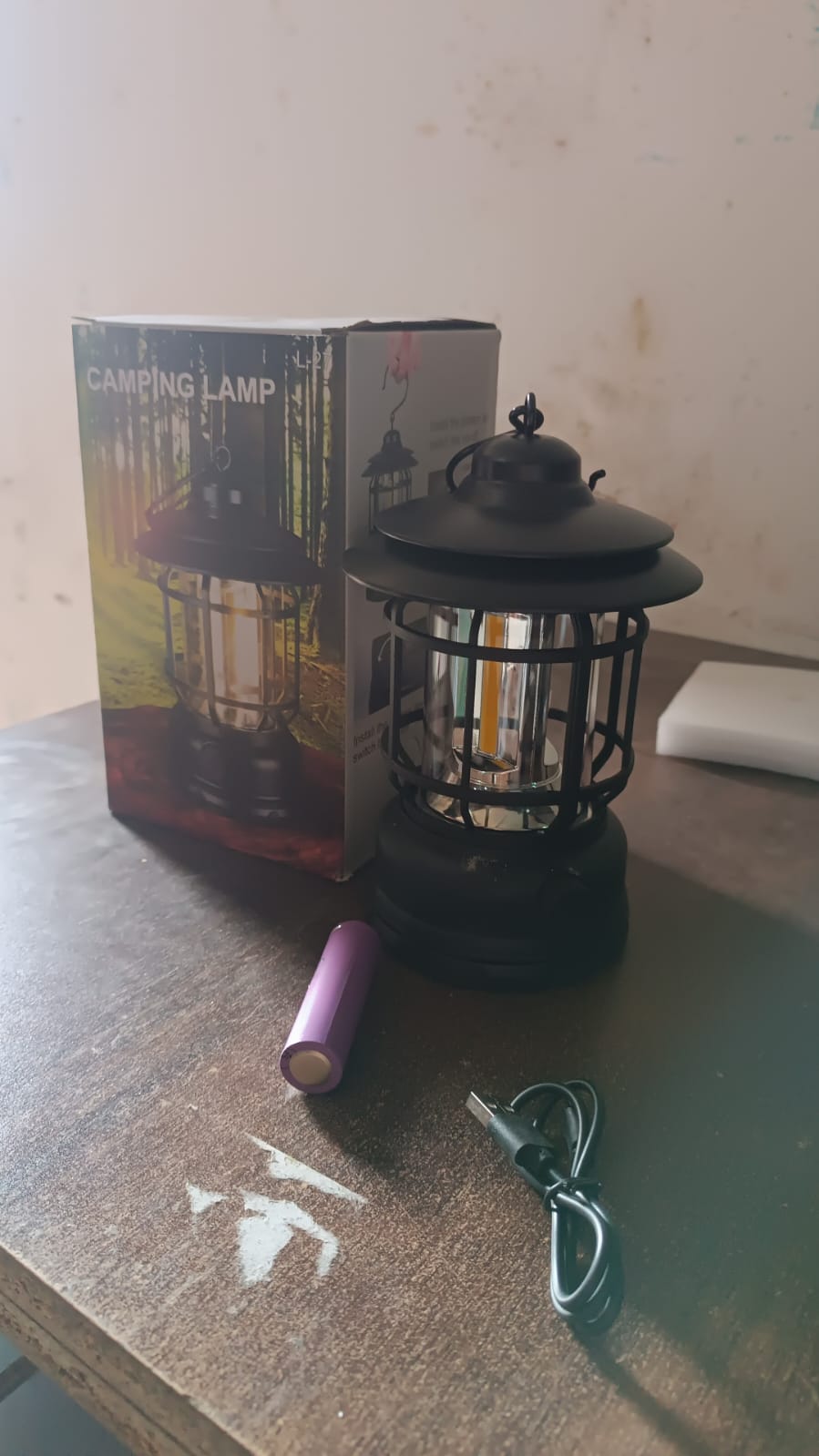 A black retro LED camping lantern, its packaging, a purple battery, and a charging cable are on a dark wooden surface.