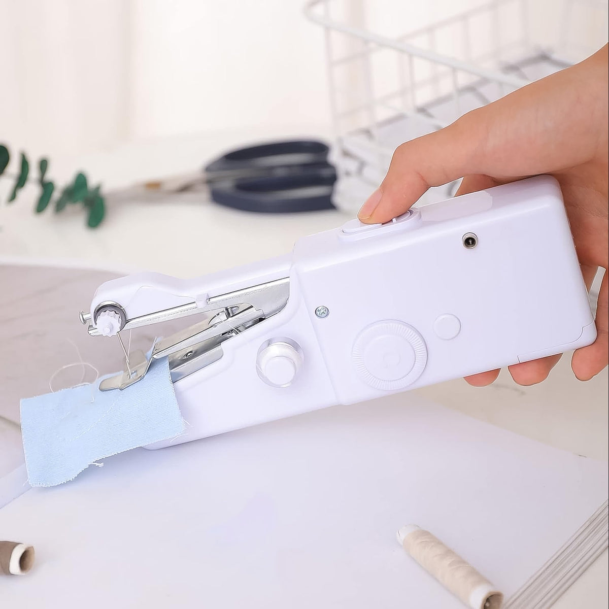 A hand holds a white handheld sewing machine stitching light blue fabric on a white desk with thread spools.