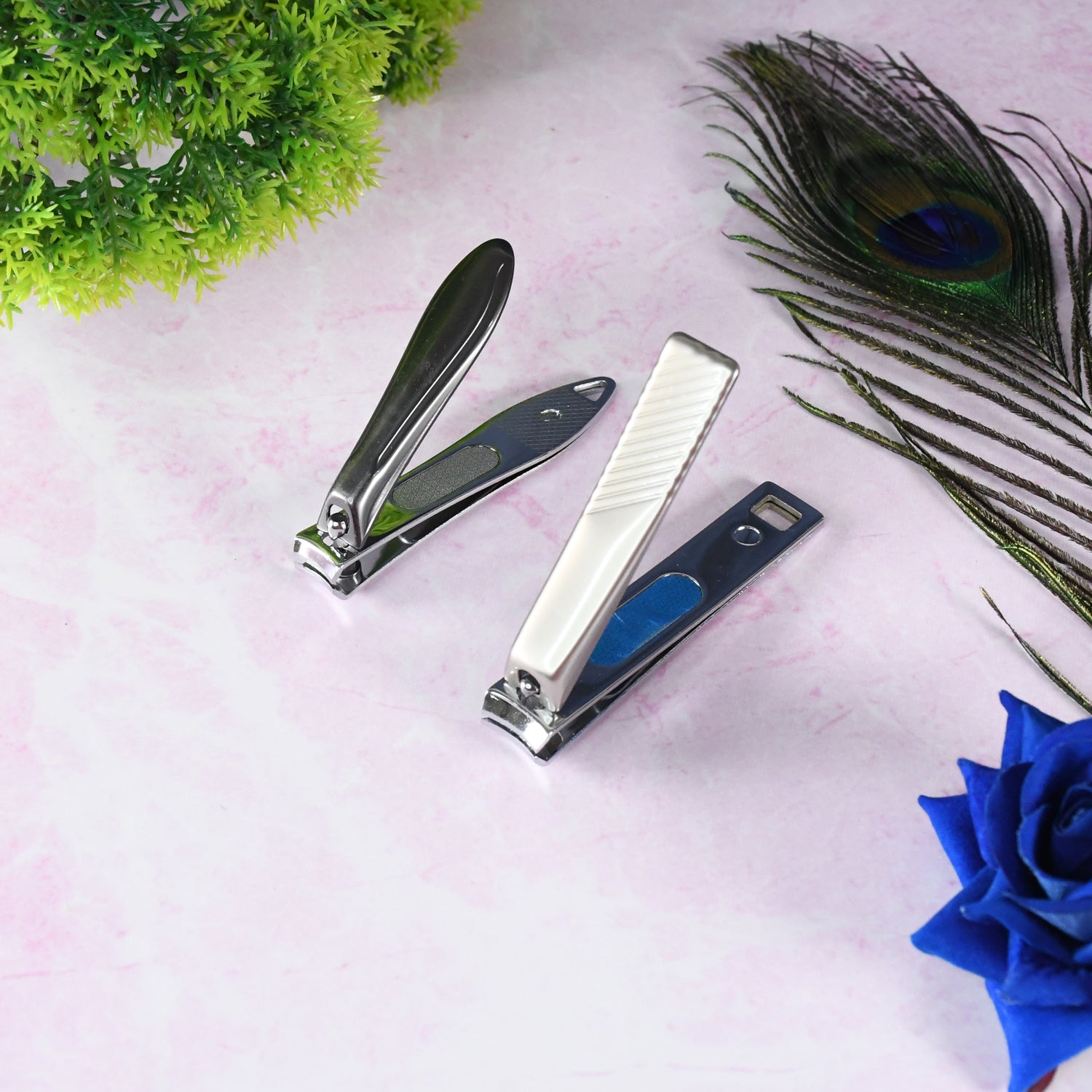 Two stainless steel nail clippers on a pink surface with a green plant and a peacock feather.