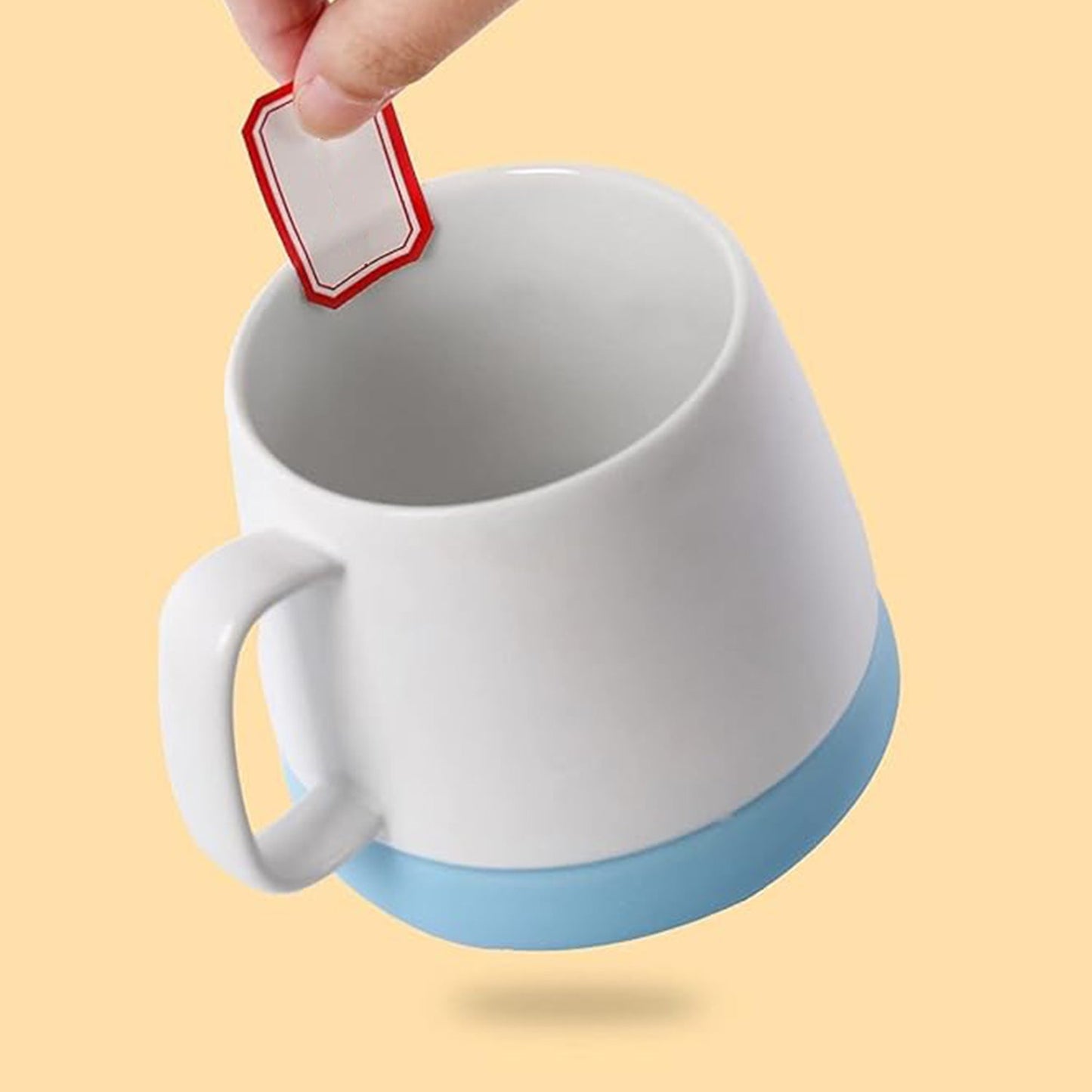 A hand peeling a red-bordered label from a sheet, above a white mug with a light blue base.