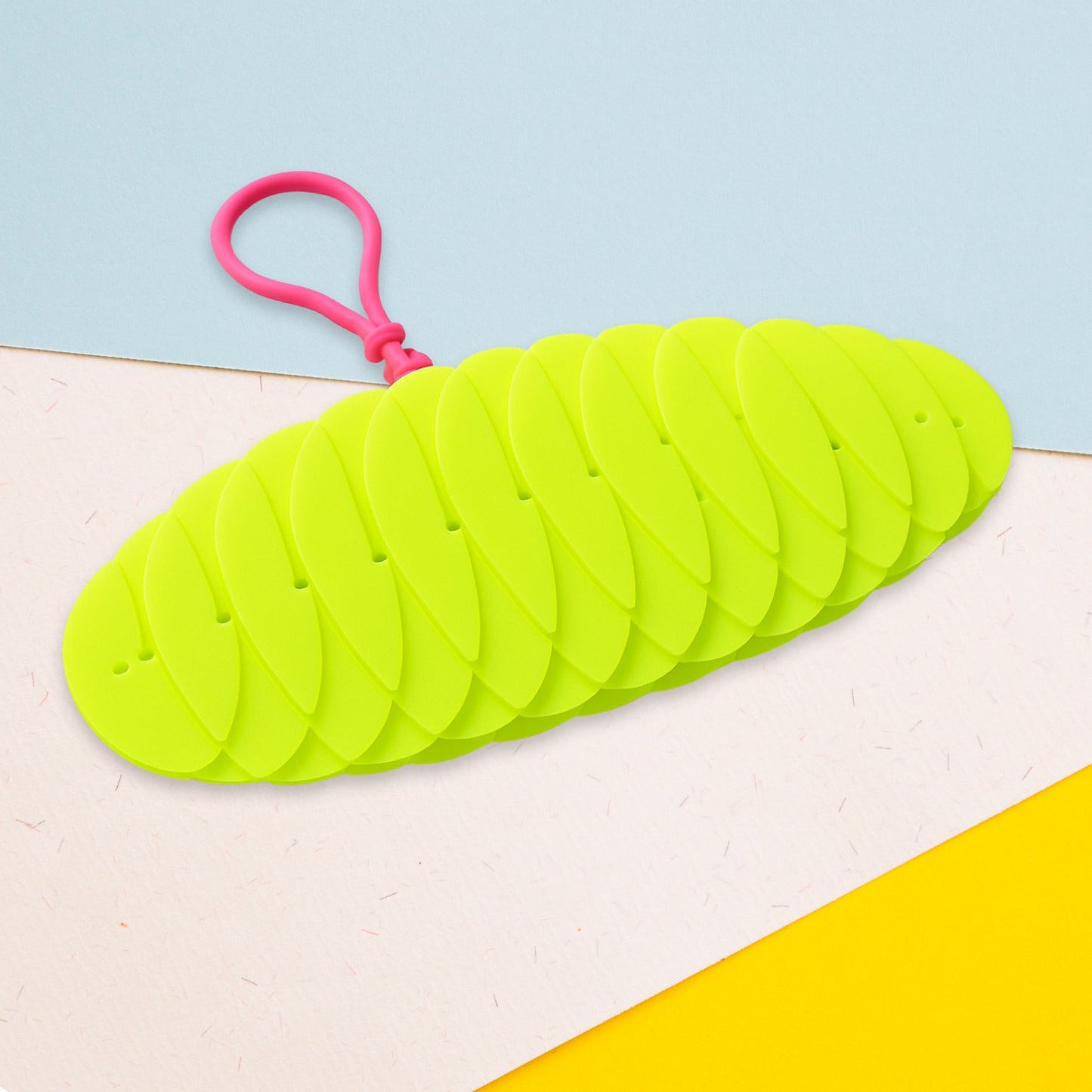 A bright green collapsible silicone ball keychain with a pink loop, shown partially collapsed on a light blue, beige, and