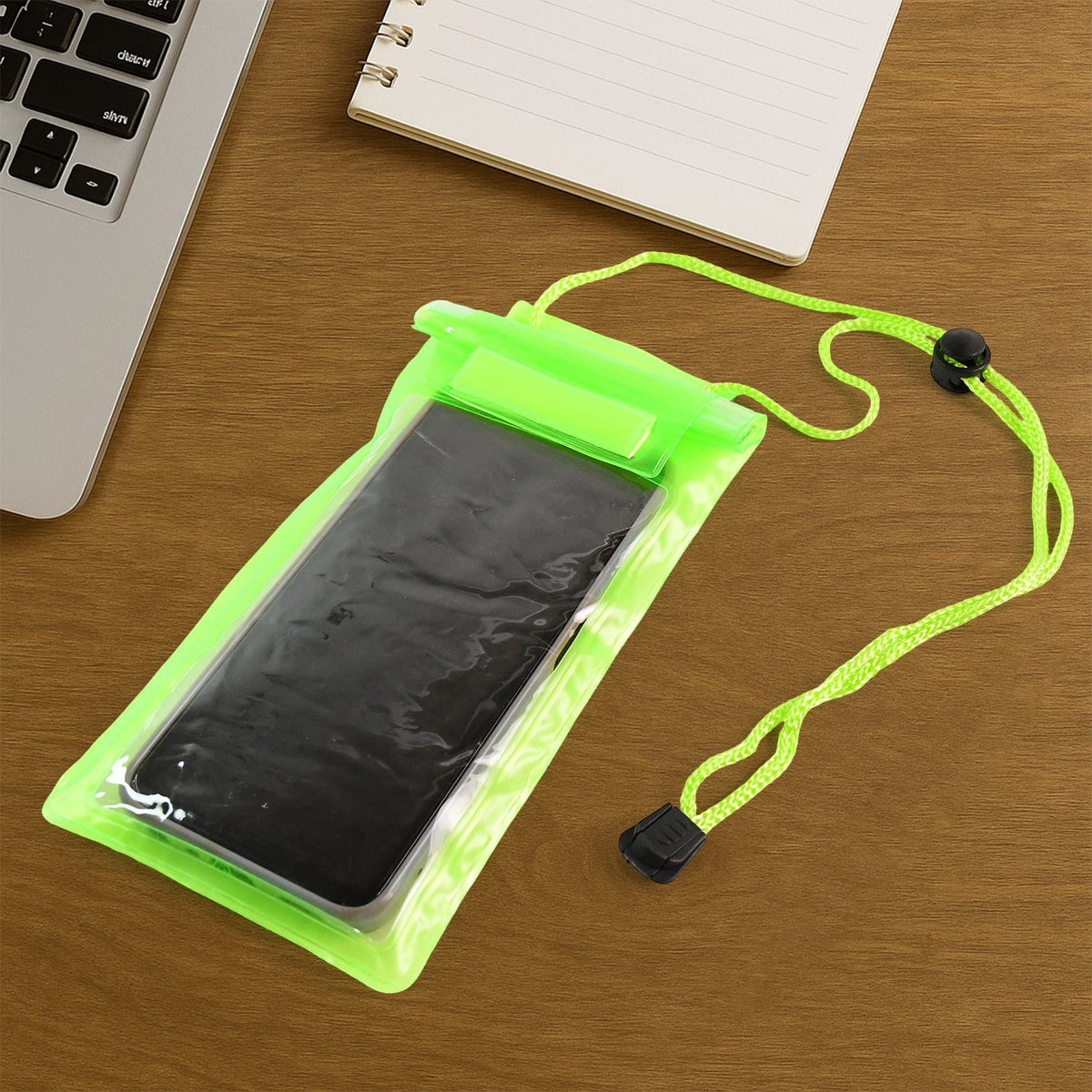 A bright green waterproof phone pouch containing a dark smartphone lies on a wooden desk next to a laptop and a notebook.