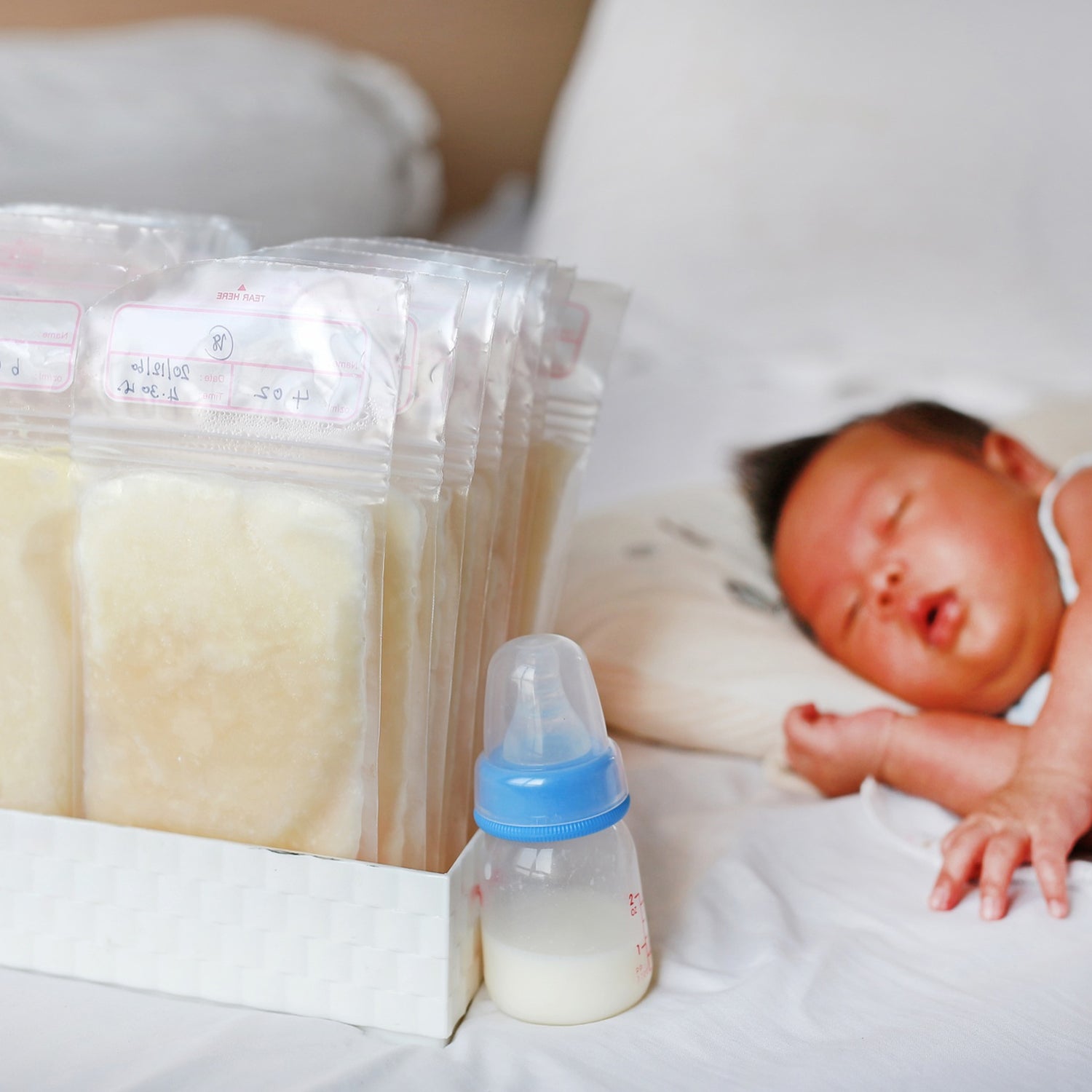 Several breastmilk storage bags filled with frozen milk stand in a container next to a sleeping baby and a baby bottle.