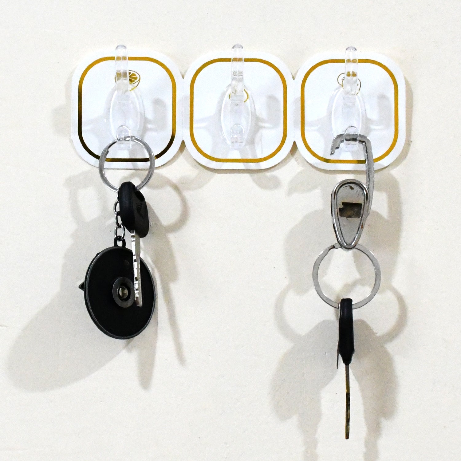 Three transparent adhesive hooks with gold borders on a white wall, holding two sets of keys.
