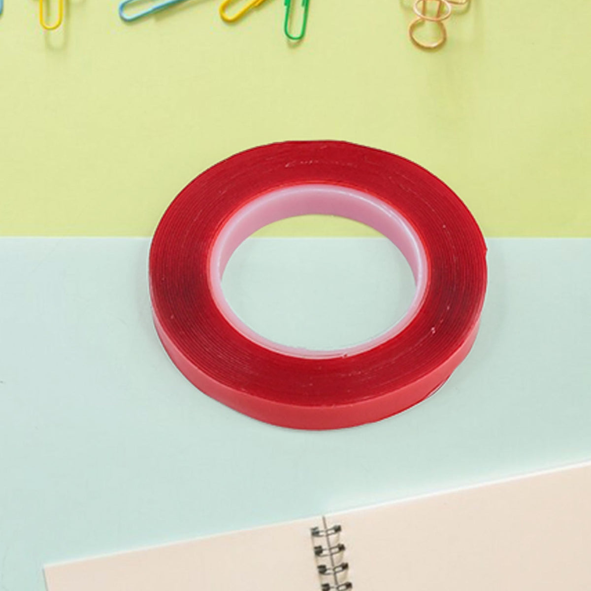 A roll of red-lined clear tape is on a light green and blue surface next to a spiral-bound notebook.