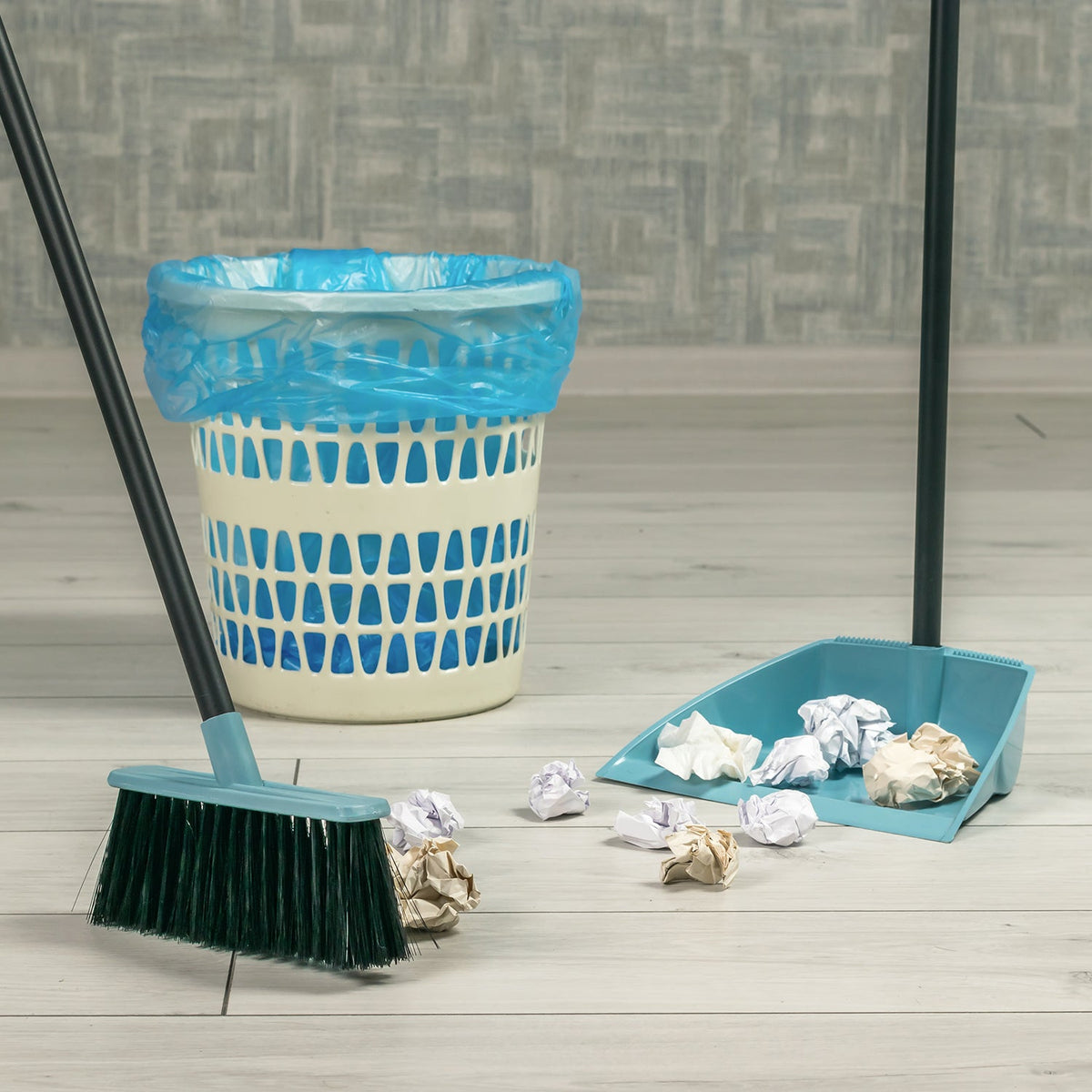 A teal broom and dustpan on a light wood floor, surrounded by crumpled paper and a white trash can with a blue liner.