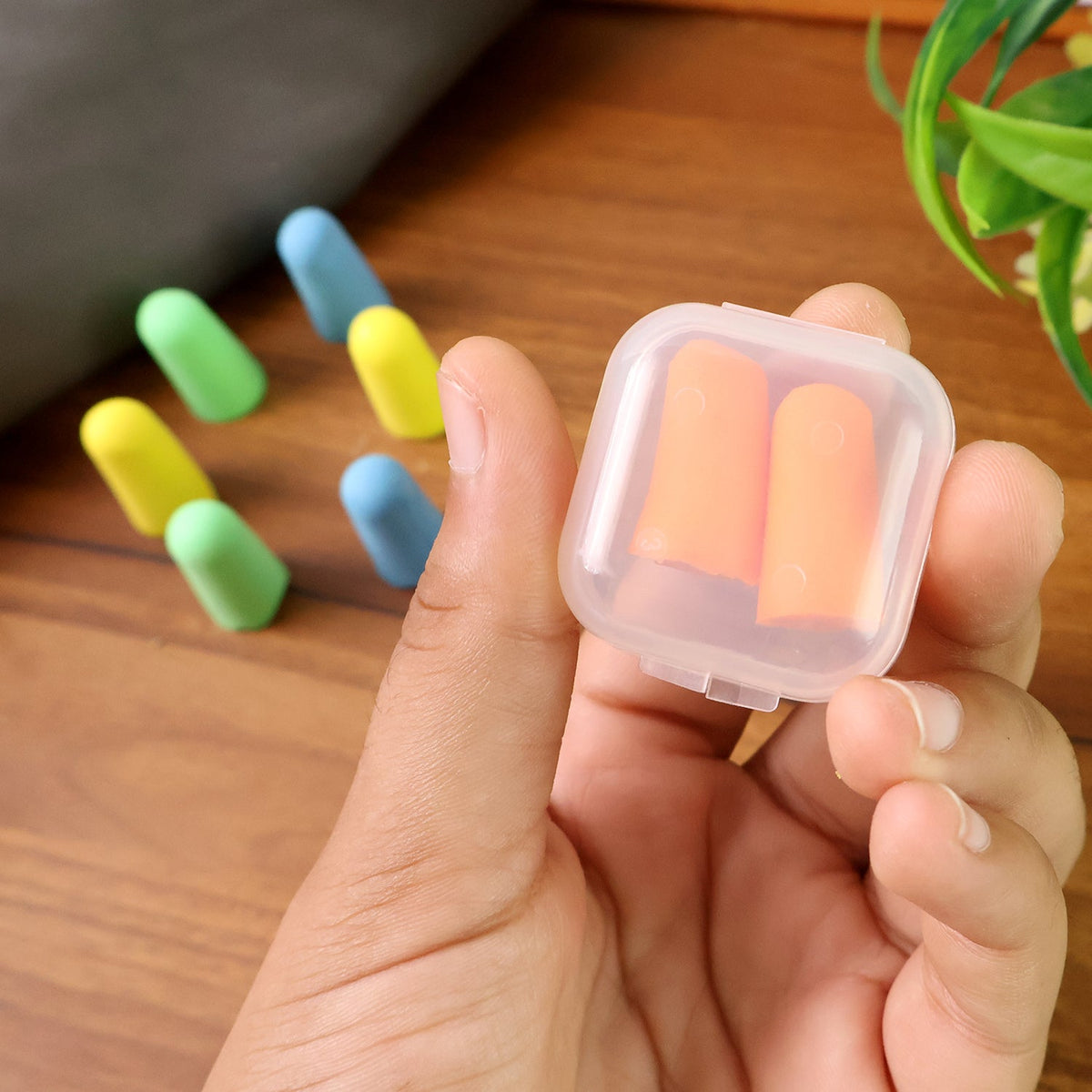 A hand holding a clear plastic case with two orange earplugs inside, with other colored earplugs blurred in the background.